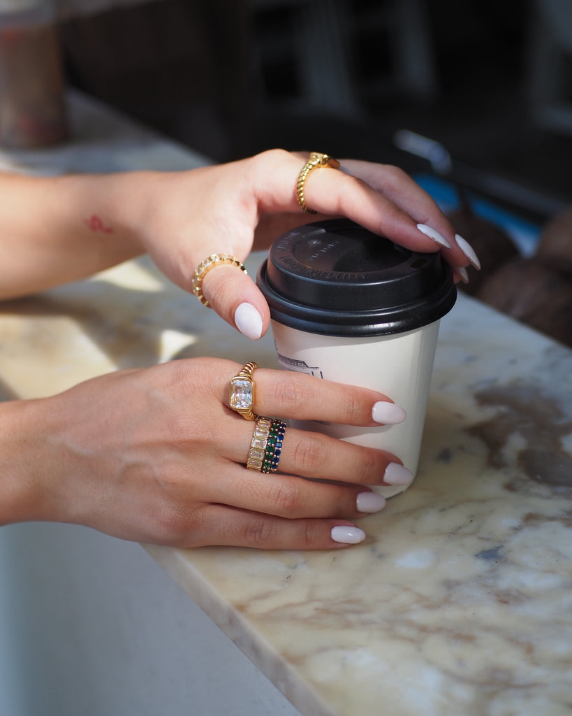 A close-up lifestyle shot of a person's hands with white manicured nails wearing gold rings, holding a white paper coffee cup with a black lid on a marble countertop.