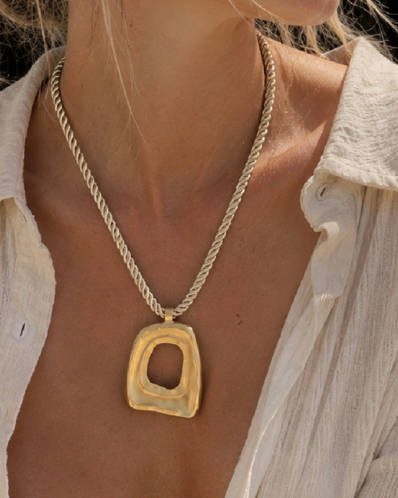 A close-up lifestyle shot of a model wearing a gold pendant necklace with a textured, abstract square-shaped pendant on a twisted chain.