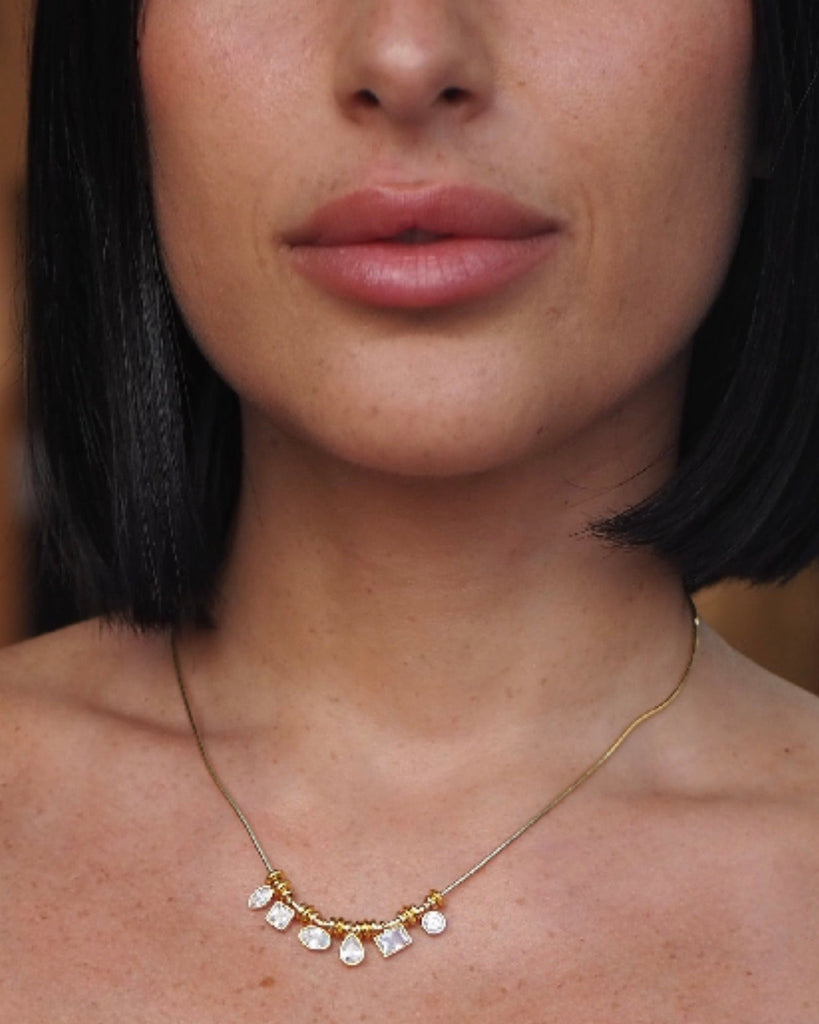 A close-up of a model wearing a delicate gold chain necklace with small white pearl-like beads and tiny clear stones at the center.