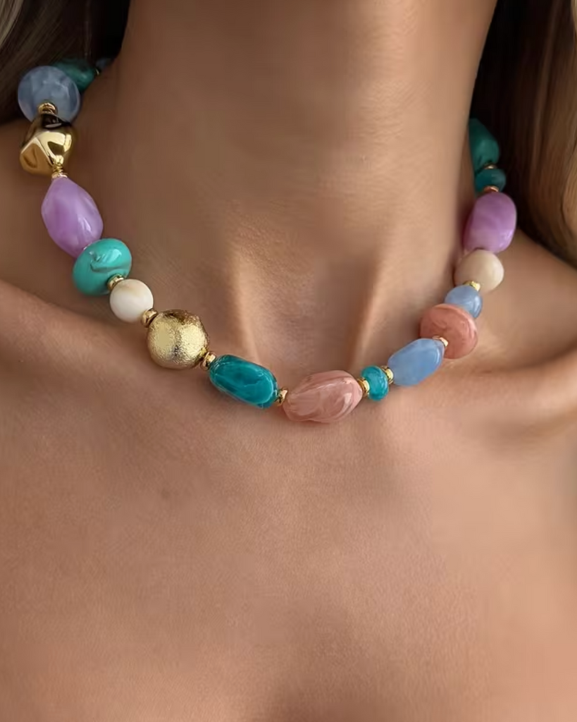 A close-up of a multicolor beaded necklace with gold-toned accents, worn on a model's neck. The necklace features pastel purple, turquoise, light blue, pink, and white beads with one prominent textured gold bead.